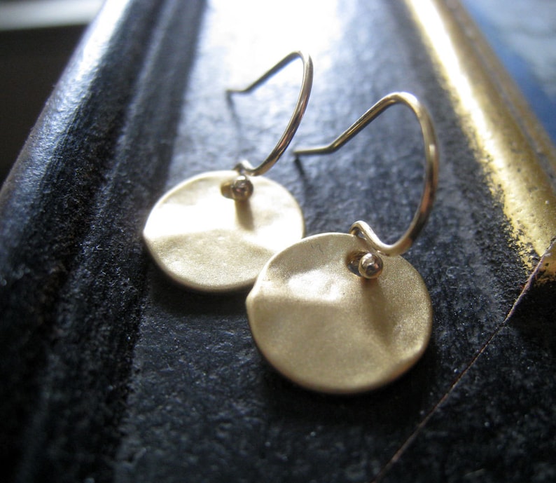 May include: A pair of gold-toned drop earrings featuring circular, hammered-texture discs suspended from thin, curved metal ear wires. Each disc is attached to the wire by a small, spherical bead, creating a simple, reflective, and metallic design.
