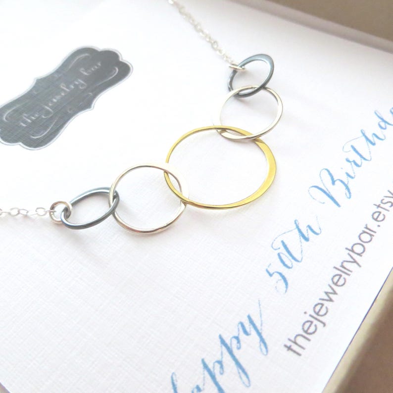 Puede incluir: Un collar plateado con cinco c&iacute;rculos entrelazados, uno dorado y los otros plateados y gris oscuro. El collar est&aacute; sobre una tarjeta blanca con la inscripci&oacute;n azul "Happy 50th Birthday". Tambi&eacute;n es visible una etiqueta gris con "the jewelry bar".