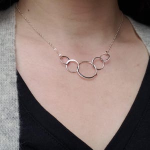 Puede incluir: Un collar plateado con cinco c&iacute;rculos abiertos interconectados de diferentes tama&ntilde;os. Los c&iacute;rculos est&aacute;n unidos por una fina cadena. El collar se muestra sobre una camiseta oscura y una rebeca gris.