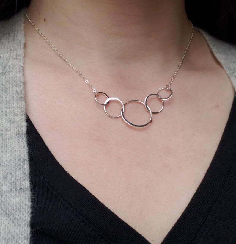 Puede incluir: Un collar plateado con cinco c&iacute;rculos abiertos interconectados de diferentes tama&ntilde;os. Los c&iacute;rculos est&aacute;n unidos por una fina cadena. El collar se muestra sobre una camiseta oscura y una rebeca gris.