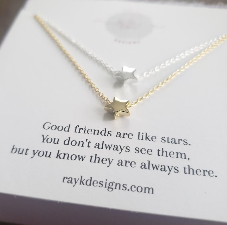 May include: Two delicate star necklaces, one gold and one silver, are displayed on a white card. The gold necklace is in the foreground, with a small star pendant. The card has text that reads, "Good friends are like stars..." and the website raykdesigns.com.