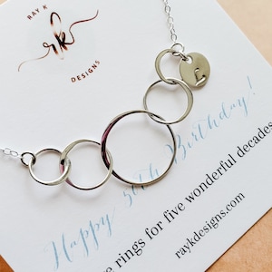 Puede incluir: Un collar plateado con cinco anillos interconectados y un peque&ntilde;o dije circular con la letra "L". El collar se exhibe en una tarjeta blanca con el texto "Happy 50th Birthday!" y "five rings for five wonderful decades".