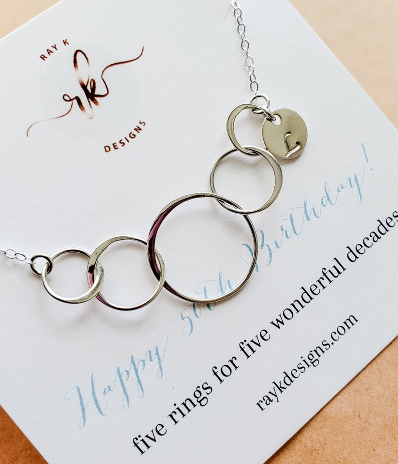 Puede incluir: Un collar plateado con cinco anillos interconectados y un peque&ntilde;o dije circular con la letra "L". El collar se exhibe en una tarjeta blanca con el texto "Happy 50th Birthday!" y "five rings for five wonderful decades".