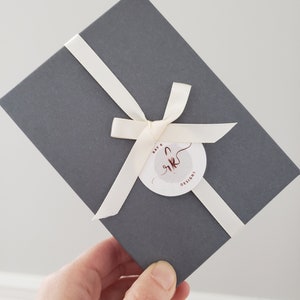 Puede incluir: Una caja de regalo gris atada con una cinta y un lazo color crema. Una etiqueta circular con el texto "Ray K Designs" est&aacute; sujeta a la cinta. La caja se sostiene en &aacute;ngulo.