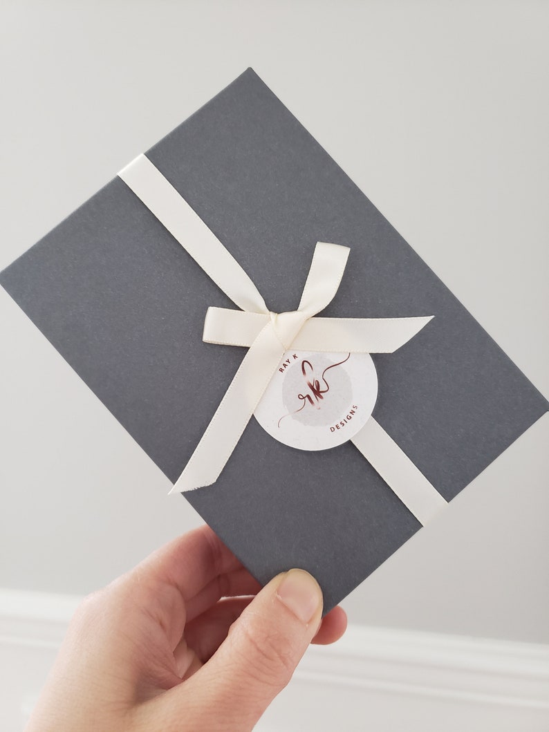Puede incluir: Una caja de regalo gris atada con una cinta y un lazo color crema. Una etiqueta circular con el texto "Ray K Designs" est&aacute; sujeta a la cinta. La caja se sostiene en &aacute;ngulo.