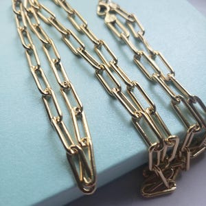 Puede incluir: Un collar de cadena dorado con eslabones rectangulares alargados. El collar se muestra sobre una superficie azul claro, mostrando su superficie brillante y reflectante. El cierre es visible, lo que indica que el collar está listo para usar.