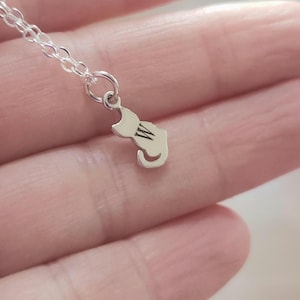 May include: A silver cat pendant necklace. The pendant is a silhouette of a cat with the letter "W" engraved on its body. The necklace chain is also silver and delicate.