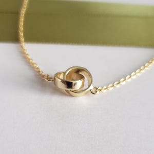 May include: A delicate gold necklace featuring a double-ring pendant. The pendant is a pair of interlocking rings, creating a knot-like design. The chain is a fine, linked style. The background includes a soft white surface and a green ribbon.