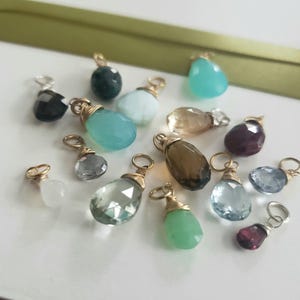 Puede incluir: Una colección de dijes de piedras preciosas en forma de lágrima, cada uno envuelto en alambre dorado con una pequeña anilla circular en la parte superior. Las piedras facetadas presentan varios colores, incluyendo transparente, azul claro, verde oscuro, marrón y morado.