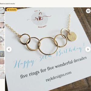 Puede incluir: Un collar dorado con cinco anillos entrelazados y un peque&ntilde;o dije redondo grabado con la letra "M". El collar se exhibe en una tarjeta con el texto "Happy 50th Birthday!" y "five rings for five wonderful decades".