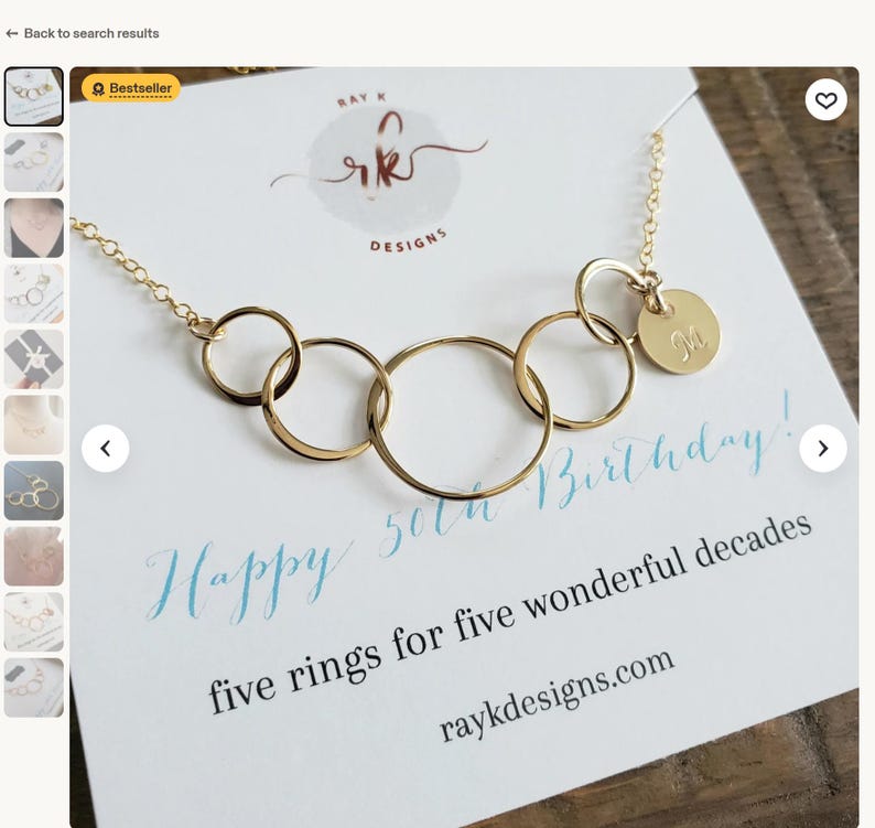 Puede incluir: Un collar dorado con cinco anillos entrelazados y un peque&ntilde;o dije redondo grabado con la letra "M". El collar se exhibe en una tarjeta con el texto "Happy 50th Birthday!" y "five rings for five wonderful decades".