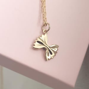 Pode incluir: Um colar com um pingente dourado em forma de gravata borboleta. O pingente está suspenso por uma delicada corrente dourada. O fundo é rosa claro.