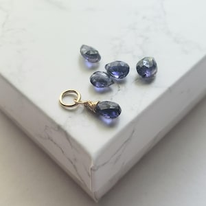 Puede incluir: Un colgante de oro con una piedra preciosa púrpura, rodeado de cinco piedras preciosas sueltas de color púrpura sobre una superficie de mármol blanco.