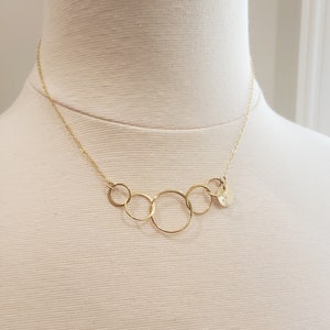 Puede incluir: Un collar dorado con cinco c&iacute;rculos interconectados y un peque&ntilde;o dije redondo. El collar se exhibe en un maniqu&iacute;, mostrando su delicado dise&ntilde;o y cadena.