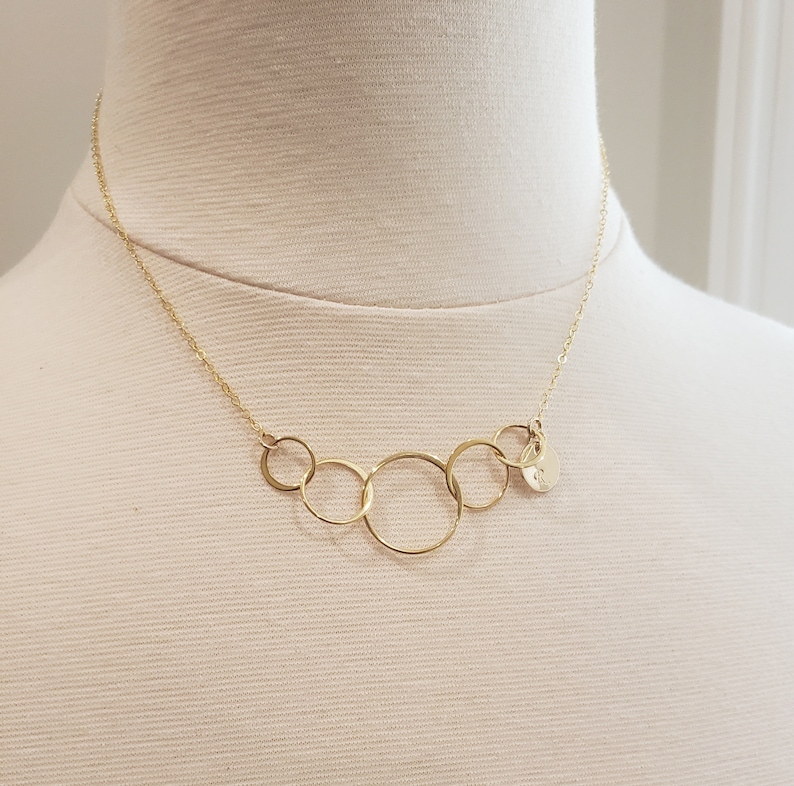 Puede incluir: Un collar dorado con cinco c&iacute;rculos interconectados y un peque&ntilde;o dije redondo. El collar se exhibe en un maniqu&iacute;, mostrando su delicado dise&ntilde;o y cadena.