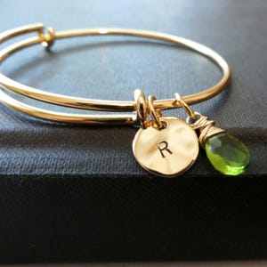 Puede incluir: Pulsera dorada con un dije redondo grabado con la letra "R" y un colgante de piedra verde. La pulsera tiene un diseño sencillo y elegante, perfecto para el uso diario. La piedra es de color verde vibrante.