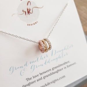 Puede incluir: Un collar de plata con tres anillos entrelazados, cada uno incrustado con pequeñas piedras brillantes. Los anillos son de oro rosa, oro y plata. El collar se muestra en una tarjeta con el texto "Abuela y Nieta".