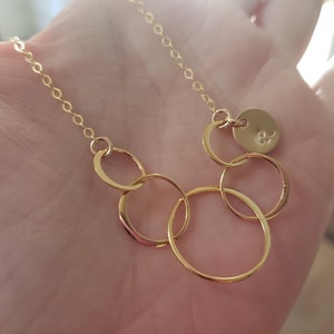 Puede incluir: Collar dorado con una cadena fina y varios c&iacute;rculos entrelazados. Un peque&ntilde;o dije redondo con una inicial grabada est&aacute; adjunto. El collar se muestra sobre un fondo suave y borroso.