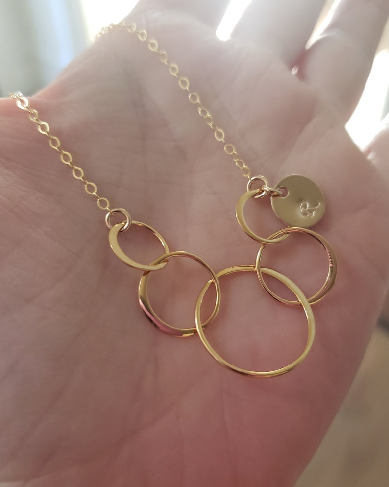 Puede incluir: Collar dorado con una cadena fina y varios c&iacute;rculos entrelazados. Un peque&ntilde;o dije redondo con una inicial grabada est&aacute; adjunto. El collar se muestra sobre un fondo suave y borroso.