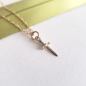 Puede incluir: Un collar con colgante dorado con un pequeño dije de daga. La daga tiene una empuñadura detallada y una hoja puntiaguda. La cadena del collar es delicada y dorada, con una cinta verde claro en el fondo.