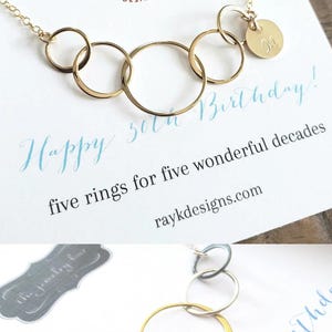 Puede incluir: Un collar dorado con cinco anillos entrelazados y un peque&ntilde;o colgante en forma de disco con la letra "M". El collar se muestra en una tarjeta con el texto "Happy 50th Birthday!" y "five rings for five wonderful decades".