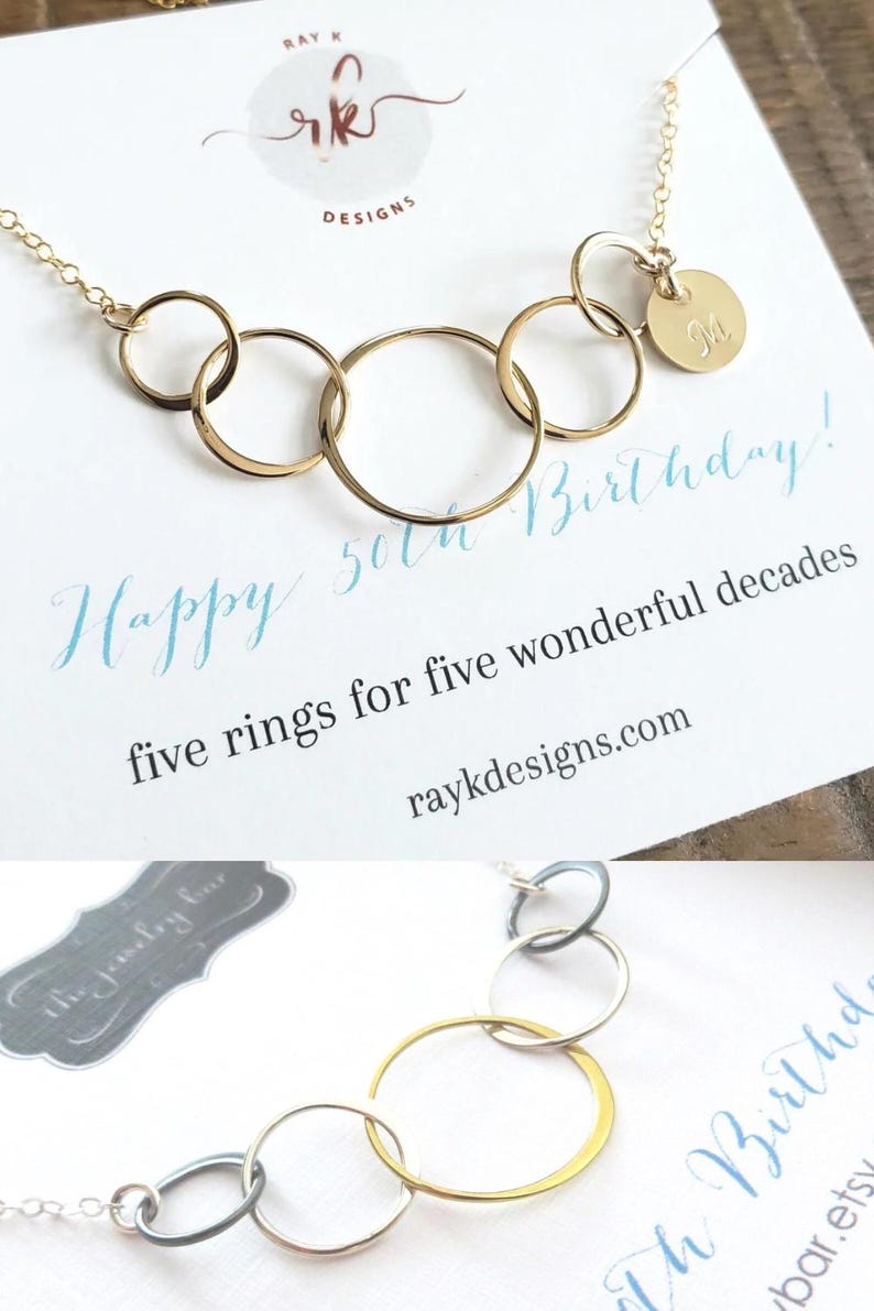 Puede incluir: Un collar dorado con cinco anillos entrelazados y un peque&ntilde;o colgante en forma de disco con la letra "M". El collar se muestra en una tarjeta con el texto "Happy 50th Birthday!" y "five rings for five wonderful decades".