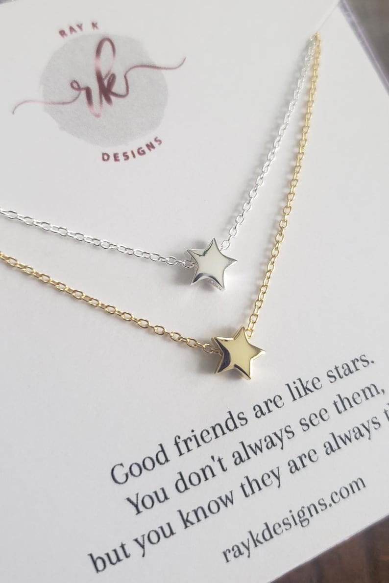 May include: Two delicate star necklaces, one silver and one gold, are displayed on a white card. The card features the text "Good friends are like stars" and the website raykdesigns.com. The necklaces are a simple, elegant design.