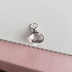 May include: A faceted, teardrop-shaped pale pink gemstone pendant secured with silver-toned wire wrapping. The gemstone features a polished surface with multiple facets that catch the light, attached to a small, circular silver-toned metal jump ring.