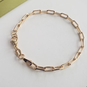 Op de afbeelding: Een goudkleurige kettingarmband met langwerpige, rechthoekige schakels. De armband is voorzien van een karabijnsluiting en een klein ovaal label met de tekst 18KGP, wat de metaalafwerking aangeeft. De schakels zijn uniform van vorm en grootte.
