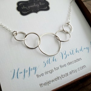 May include: A silver necklace with five interlocking circles, a gift for a 50th birthday. The necklace is displayed in a white box with the text "Happy 50th Birthday! five rings for five decades thejewelrybar.etsy.com"