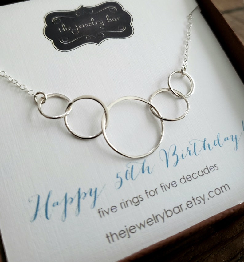 May include: A silver necklace with five interlocking circles, a gift for a 50th birthday. The necklace is displayed in a white box with the text "Happy 50th Birthday! five rings for five decades thejewelrybar.etsy.com"
