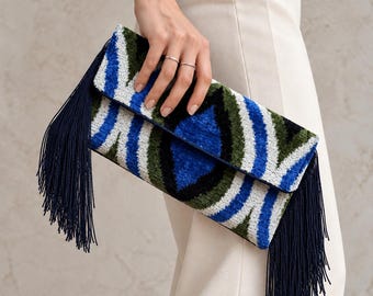 Könnte beinhalten: Eine rechteckige Clutch mit einem lebendigen Samtmuster in Königsblau, Waldgrün, Schwarz und Weiß. Das Accessoire ist mit langen, dunkel marineblauen Fransen versehen, die an beiden Seiten der Umschlagklappe herabhängen.