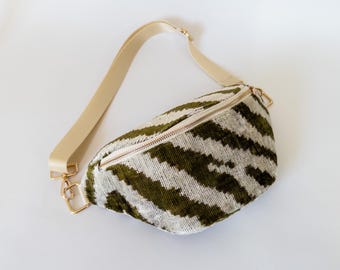 May include: A green and white striped bum bag with a beige strap and gold-coloured hardware. The bag has a zip closure and a soft, textured appearance. The strap is adjustable.