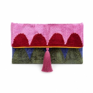 Op de afbeelding: Een rechthoekige clutch met een roze bovenkant met drie bordeauxrode bergachtige vormen. Het onderste gedeelte is groen en blauw met een gele rand. Een roze kwast hangt aan de onderkant.