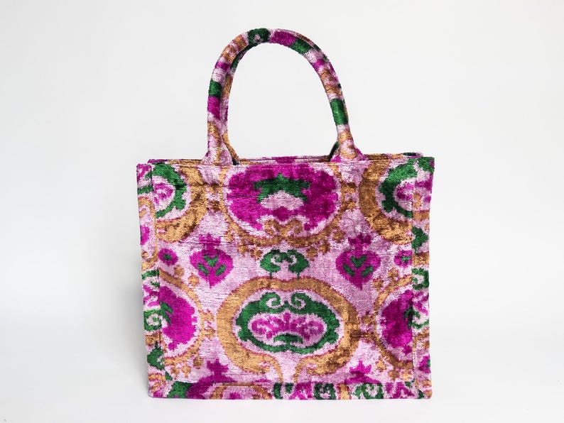May include: A pink, green, and gold floral tote bag with a velvet texture. The bag has a top handle and a spacious interior.