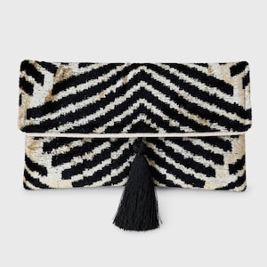 Puede incluir: Un bolso de mano rectangular con un patrón de chevrón en negro y crema con una textura aterciopelada. El bolso presenta un diseño de solapa plegable y está adornado con una borla negra única que cuelga del centro del borde frontal.
