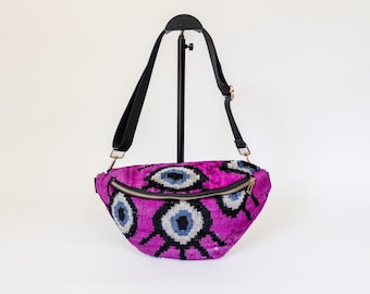 May include: A fuchsia fanny pack featuring a repeating eye design in blue, grey, and black. It has a black adjustable strap and a gold zip. The bag is displayed on a black stand against a white backdrop.