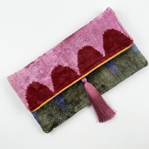 Peut inclure: Une pochette rectangulaire en velours avec un motif festonné rose et rouge foncé sur le rabat. Le corps principal présente un motif vert foncé et bleu. Un pompon rose est suspendu au centre du bord inférieur du rabat.