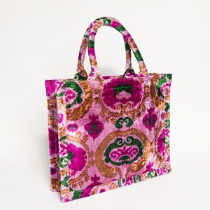 May include: A pink, gold, and green floral tote bag with a velvet texture. The bag has two top handles.