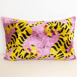 May include: A rectangular cushion featuring a pink background and a repeating pattern of yellow tigers with black stripes. The cushion is made of a soft, velvety material.