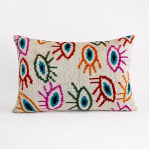 May include: A rectangular throw pillow with a cream-colored base featuring a repeating pattern of stylized eye motifs in vibrant colors including red, orange, pink, dark green, and bright blue. The eyes have distinct lashes and dark pupils.