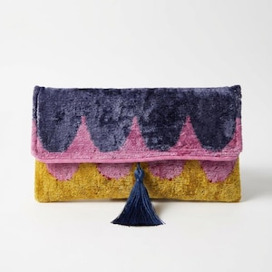 Può includere: Una pochette rettangolare in velluto con un motivo a festoni nei colori viola scuro, rosa acceso e giallo senape. La patta di chiusura è decorata con una singola nappa in filo blu scuro appesa al centro del bordo festonato.