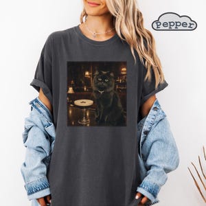 Könnte beinhalten: Ein anthrazitfarbenes T-Shirt mit einem Grafikdruck einer flauschigen schwarzen Katze mit Halsband, die an einer Bar neben einem Cocktailglas sitzt. Das Wort pepper ist in einem wolkenförmigen Logo oben rechts im Bild aufgedruckt.