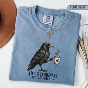 Könnte beinhalten: Ein kurzärmeliges T-Shirt in Jeansblau mit einer Grafik eines schwarzen Raben, der eine dampfende Tasse hält. Der Text unter dem Raben lautet, Hello Darkness My Old Friend. Das T-Shirt hat ein Comfort Colors Markenetikett am Innenkragen.