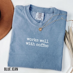 May include: A blue jean coloured t-shirt with a crew neckline and the white text "works well with coffee" printed on the chest. The shirt features a Comfort Colors brand label at the inner neck and is paired with a gold-toned pendant necklace.