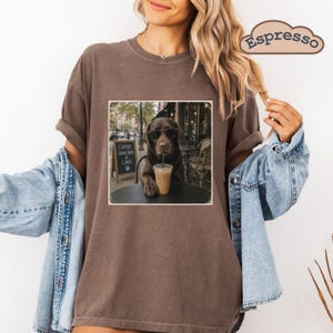 May include: A brown t-shirt featuring a graphic of a chocolate Labrador wearing sunglasses, sitting at a cafe table with an iced coffee. The graphic includes a chalkboard sign that reads Coffee Good Days & Labs. A light blue denim jacket is layered over.