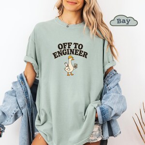 Może przedstawiać: Szałwiowy t-shirt z okrągłym dekoltem, ozdobiony grafiką przedstawiającą gęś w żółtym kasku ochronnym, trzymającą plan budowy i kalkulator. Nad grafiką znajduje się napis "OFF TO ENGINEER" wykonany pogrubioną czarną czcionką. Krótkie rękawy.