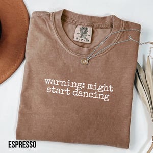 Puede incluir: Camiseta marrón con la frase "warning: might start dancing" en blanco. Un collar plateado con un pequeño colgante descansa sobre la camiseta. La camiseta está doblada y tiene una etiqueta en el cuello.