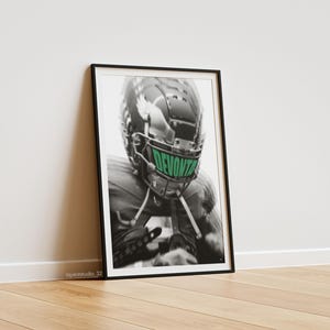Puede incluir: Impresión en blanco y negro enmarcada que presenta a un jugador de fútbol americano con un casco con el nombre "DEVONTE" en verde en la visera. La impresión está apoyada contra una pared blanca sobre un suelo de madera.