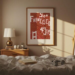May include: A framed print featuring the words "San Francisco 49ers" in red and white. The artwork hangs above a wooden bedside table with a lamp, books, and headphones on a bed with white bedding. A mirror leans against the wall.